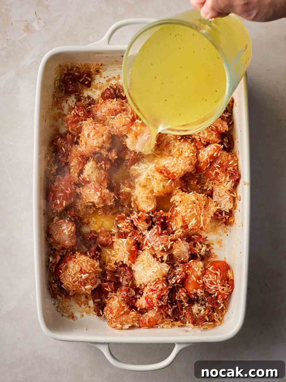 Marry Me Chicken and Rice — One-Pan Dump and Bake Recipe 6 A jug of stock being poured over an oven dish with chicken, tomatoes and rice in.
