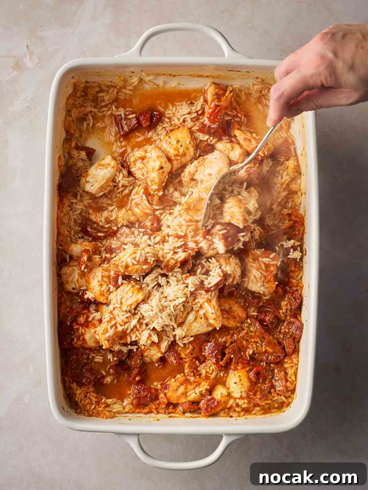 Marry Me Chicken and Rice — One-Pan Dump and Bake Recipe 7 A hand with spoon stirring an oven dish fulled with chicken, rice and stock.