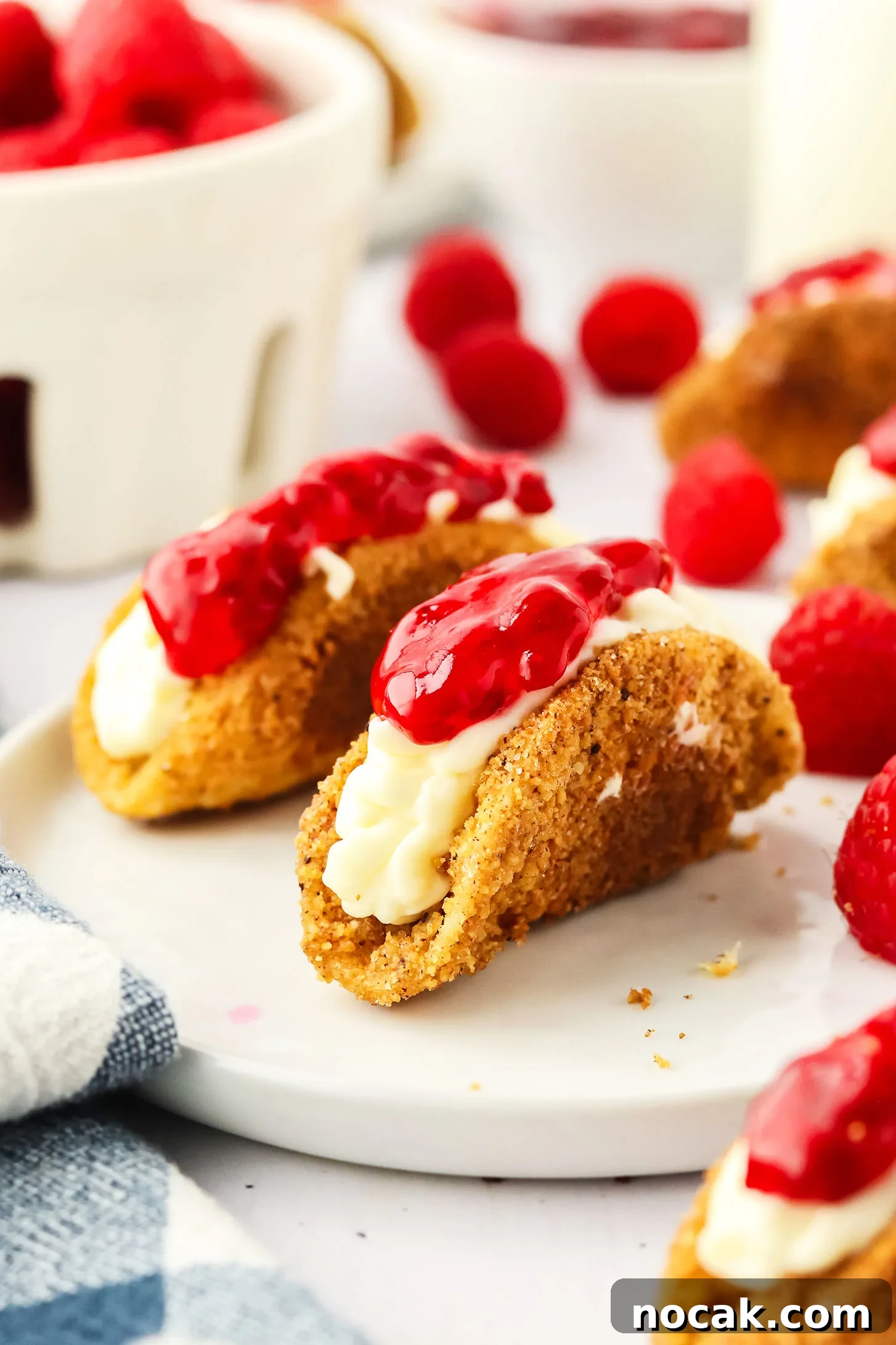 Raspberry Cheesecake Tacos: Creamy Dessert Taco Recipe 12 cheesecake tacos on a plate surrounded by fresh raspberries and a blue and white napkin