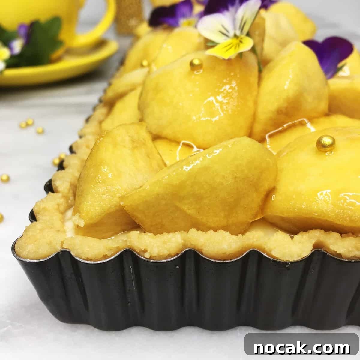 Apple tart decorated with gold pearls and flours in a tart pan with a yellow cup on the background.
