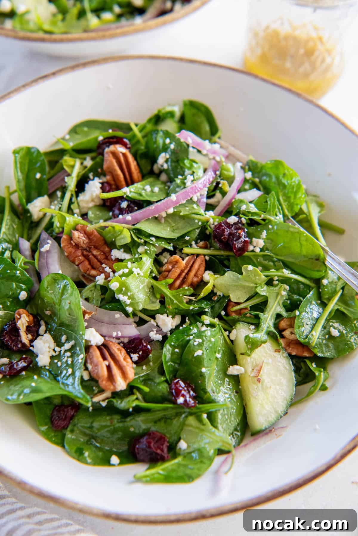A light and refreshing spinach and arugula salad sits in a white bowl for serving. The spinach and arugula is topped with cucumber, dried cranberries, red onion, pecans, feta cheese and dressing.