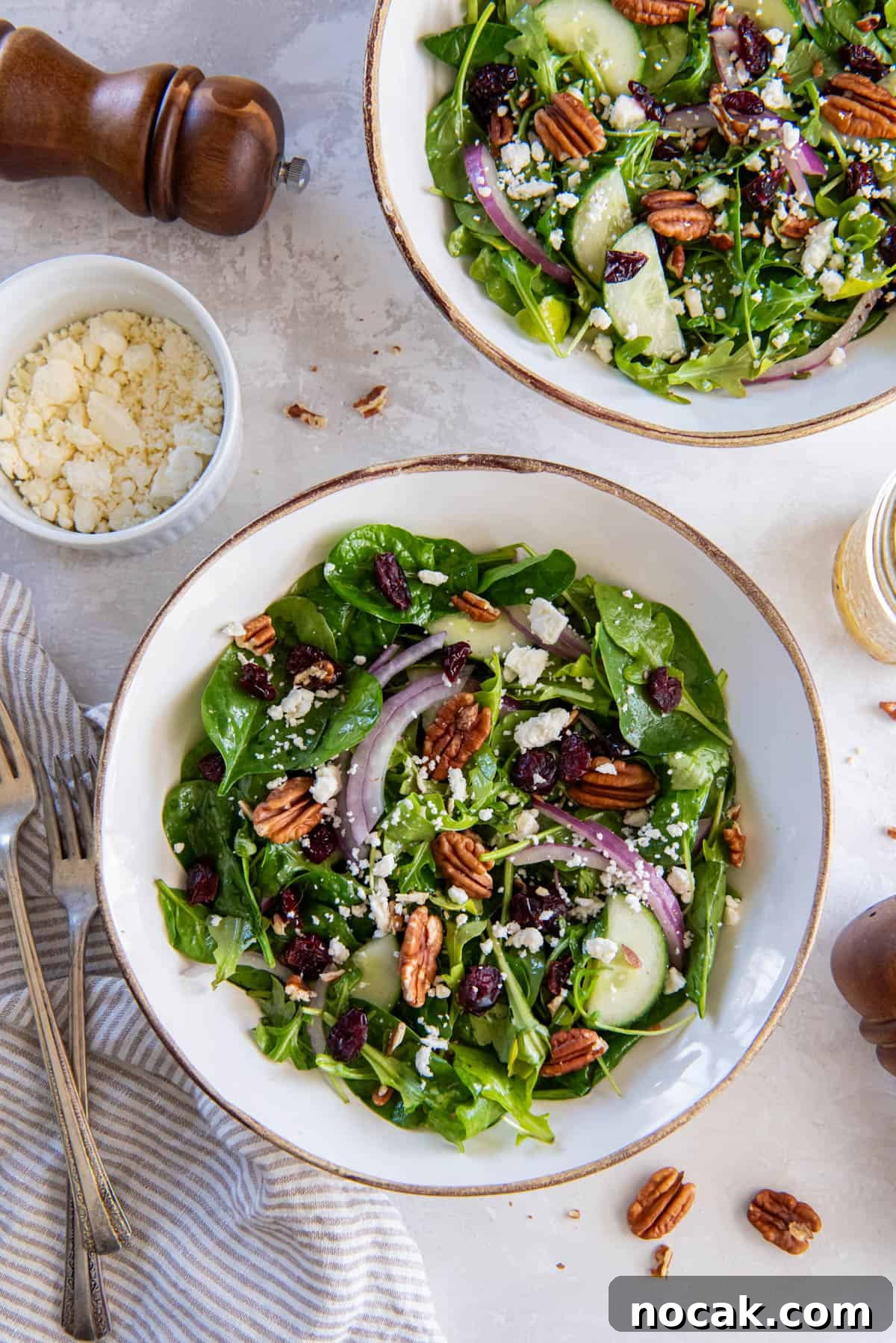 Two bowls filled with spinach arugula salad on a white tabletop for serving. A bowl of crumbled feta and cracked black pepper sits to the side of the salad bowls.
