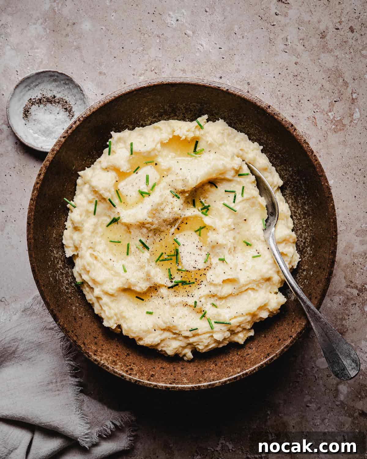 Creamy Horseradish Mashed Potatoes Recipe for Flavorful Sides 11 A rustic brown bowl filled with creamy mashed potatoes, topped with butter, chopped chives, and black pepper. A spoon rests in the bowl, with a small dish of salt nearby on a textured surface.