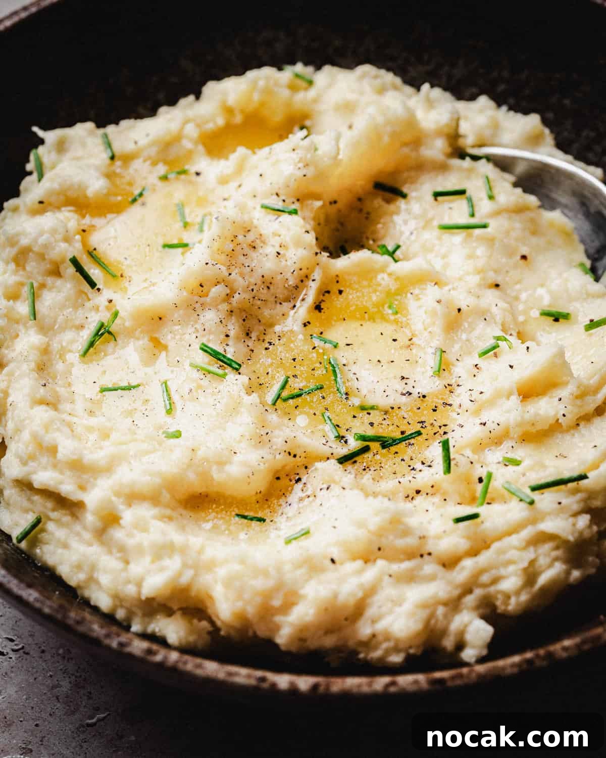 Creamy Horseradish Mashed Potatoes Recipe for Flavorful Sides 12 A bowl of creamy mashed potatoes topped with melted butter, chopped chives, and a sprinkle of black pepper, with a spoon resting in the dish.