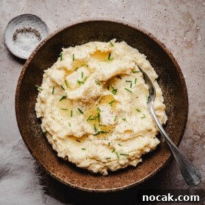 Creamy Horseradish Mashed Potatoes Recipe for Flavorful Sides 13 A rustic brown bowl filled with creamy horseradish mashed potatoes, topped with chopped chives, ground black pepper, and a drizzle of melted butter, with a spoon resting inside. A small dish of salt sits nearby on a textured surface.
