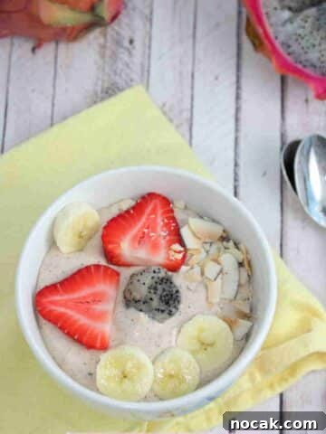 White bowl with a dragon fruit smoothie bowl in it topped with bananas, strawberries, and coconut on a yellow linen with berries, dragon fruit, and spoons around it.