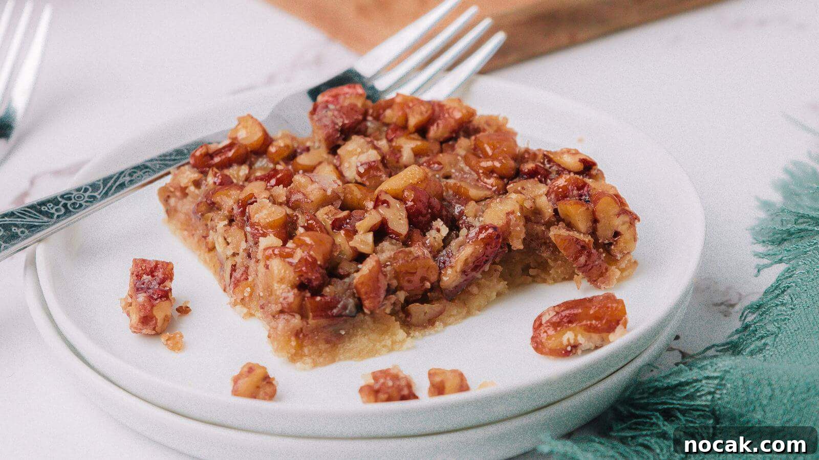 30 Christmas Desserts That Steal the Spotlight from Cookies 3 A piece of pecan bar on a white plate, with a fork resting on the side. The bar has a nutty topping and a crumbly base, partially eaten. Two forks are in the background.