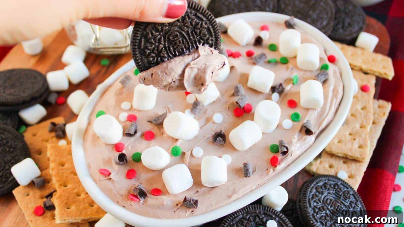 30 Christmas Desserts That Steal the Spotlight from Cookies 32 A hand holds a cookie dipped in creamy chocolate spread topped with marshmallows, chocolate chips, and red and green sprinkles. Surrounded by cookies and graham crackers on a platter.