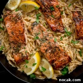 Square image of pan-fried salmon fillets nestled in a bed of creamy lemon orzo topped with parsley. There are lemon wedges at the edges of the pan.