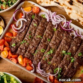 A tray of Sheet Pan Kebabs garnished with chopped parsley, red onion, and cherry tomato halves, served with flatbreads and salad on a wooden table.