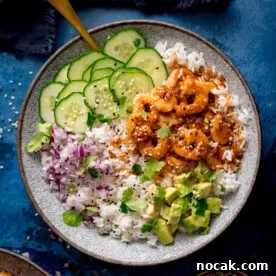 A bowl of white rice topped with cooked honey garlic prawns, sliced cucumber, chopped red onion, diced avocado, fresh coriander, and sesame seeds.