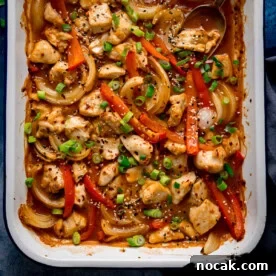 Overhead of a chicken 'stir fry' traybake with diced chicken, sliced red peppers, onions, spring onions, sesame seeds and sauce in a white dish.