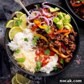 A minced beef fajita bowl with white rice, grated cheese, seasoned minced beef with peppers, sliced red onions, avocado, lime wedges, soured cream, salsa, and coriander.