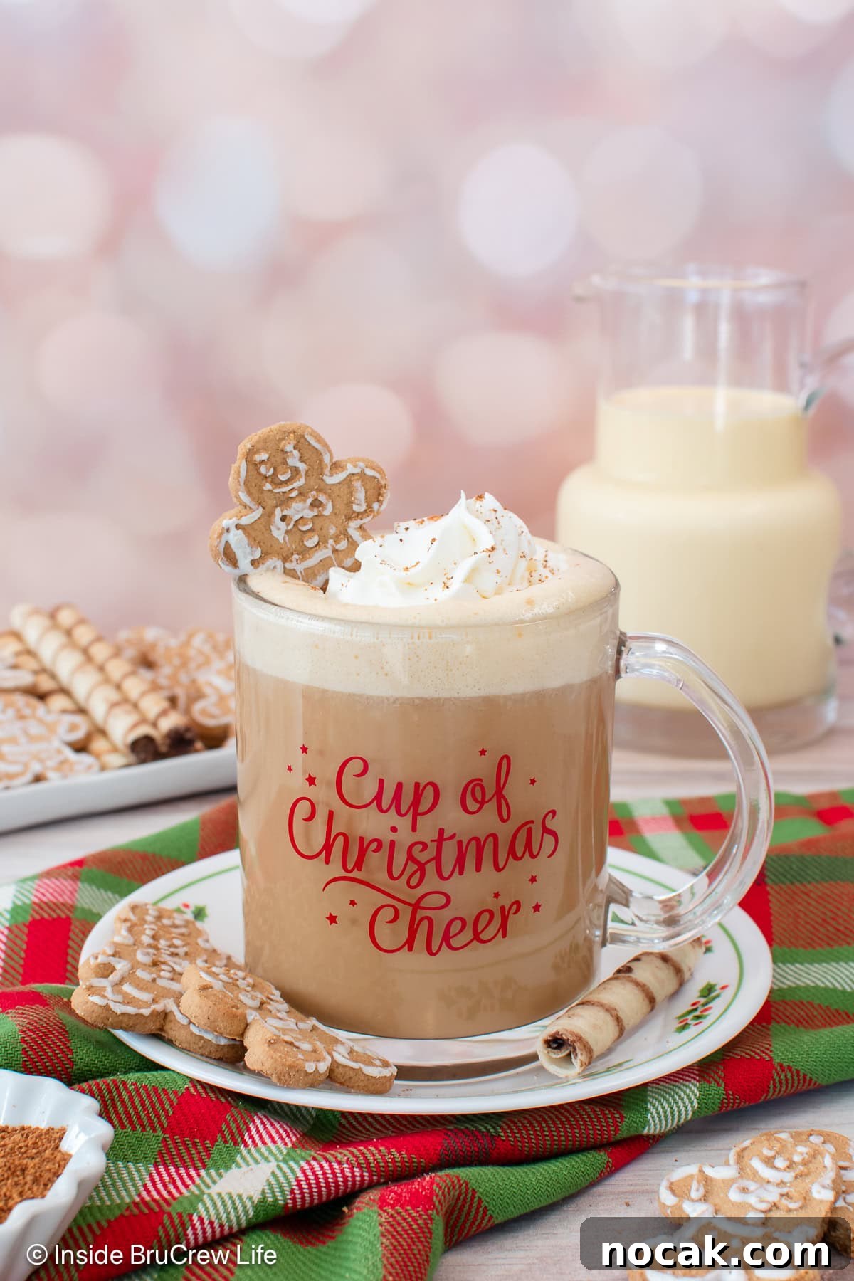 An eggnog latte topped with a swirl of whipped cream and a mini gingerbread man in a festive mug, set on top of a red and green plaid cloth.