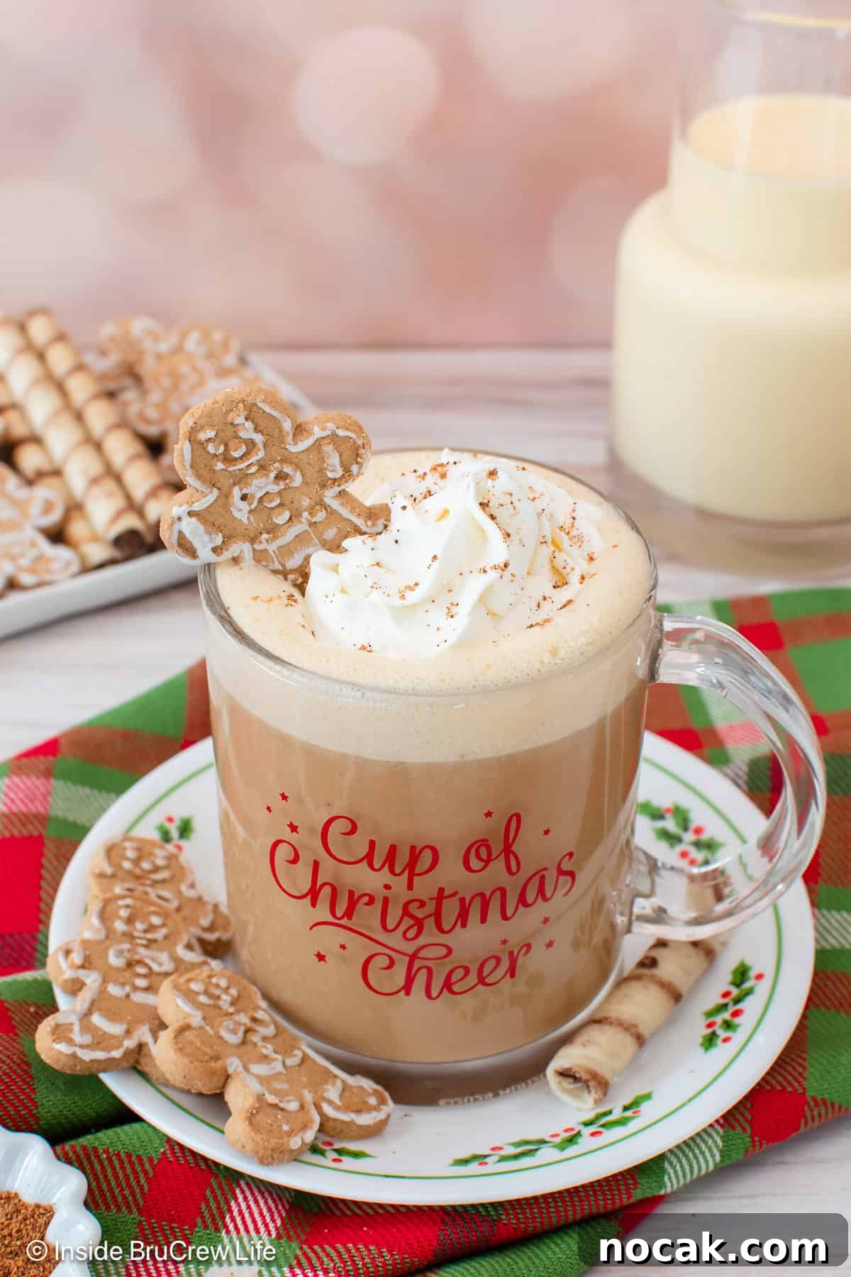 An eggnog latte topped with a swirl of whipped cream and a mini gingerbread man in a festive mug, set on top of a red and green plaid cloth.