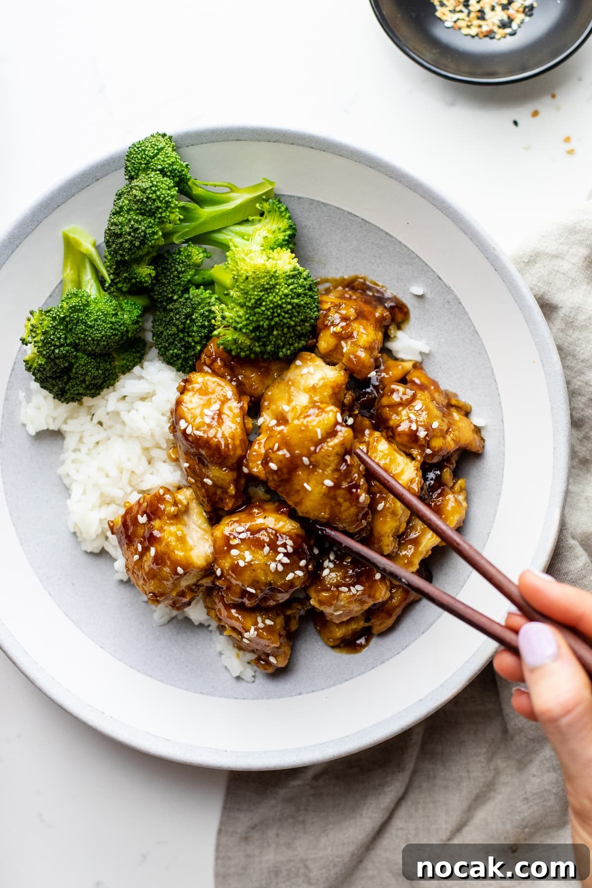 A ceramic bowl with white rice, broccoli, and honey sesame chicken with chop sticks picking up a piece of chicken.