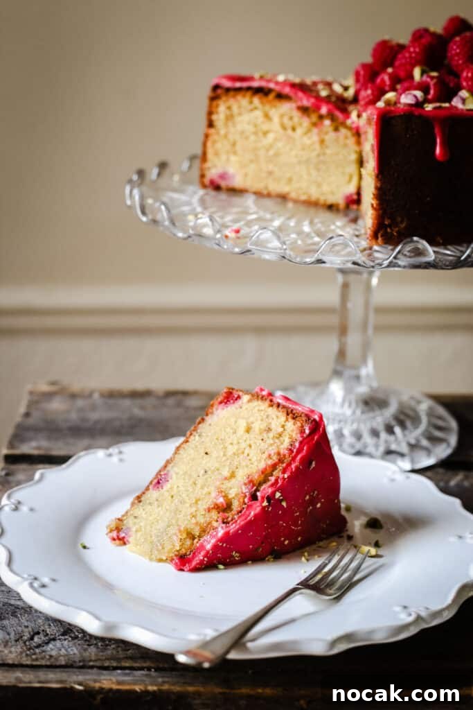 Gluten-Free Raspberry and Pistachio Cake Recipe 6 A slice of Raspberry Pistachio Cake on a white plate with a fork on a wooden table in front of a cake stand with cake on it