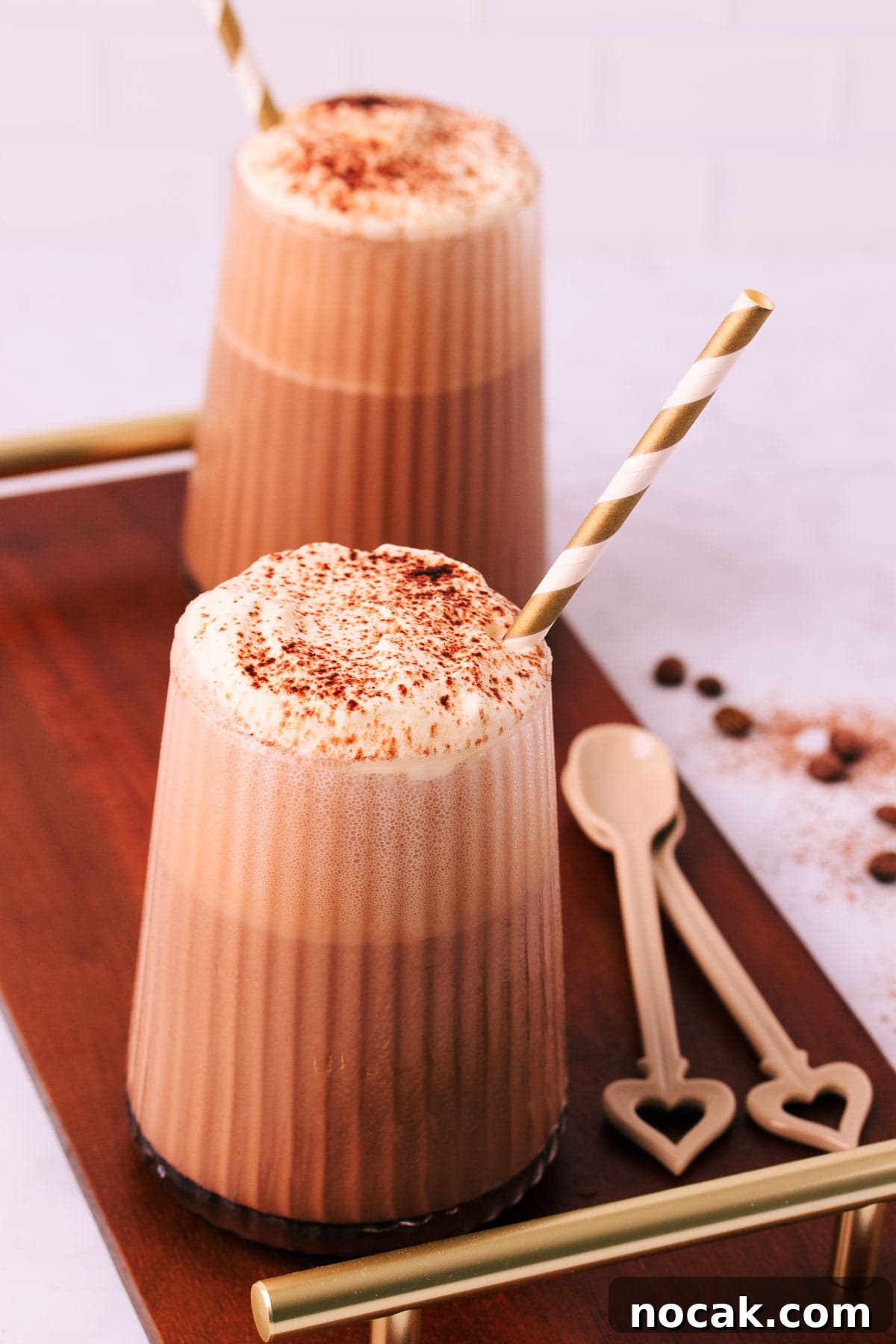 Creamy Mocha Milkshake Recipe: How to Make the Perfect Coffee Chocolate Shake 2 Two mocha milkshakes in glasses with a ribbed texture, topped with whipped cream and a sprinkle of cocoa, with gold and white striped paper straws on a dark wooden board with gold handles.