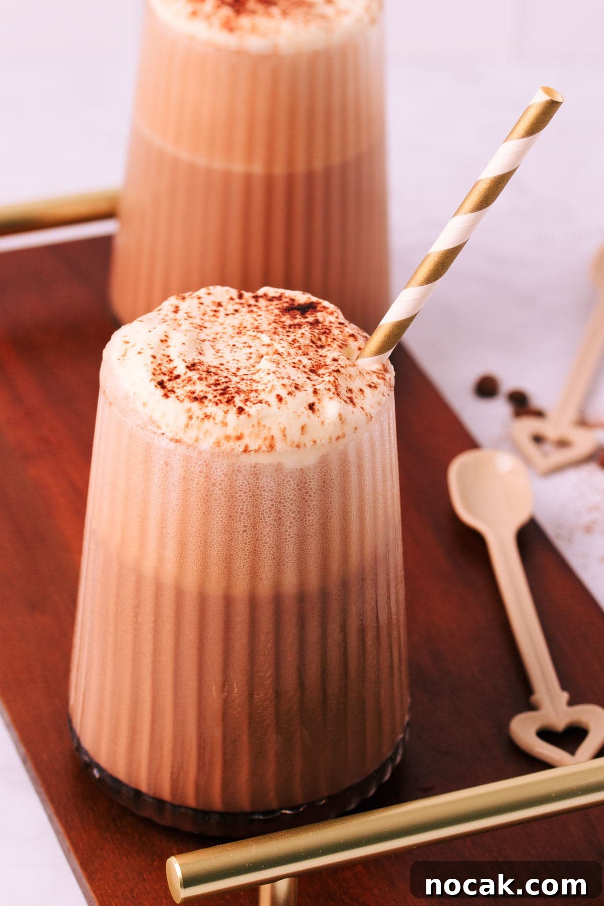 Creamy Mocha Milkshake Recipe: How to Make the Perfect Coffee Chocolate Shake 3 A mocha milkshake in a tapered glass with a ribbed texture, topped with whipped cream and a sprinkle of cocoa, with a gold and white striped paper straw on a dark wooden board with gold handles and a mocha-coloured ceramic spoon.