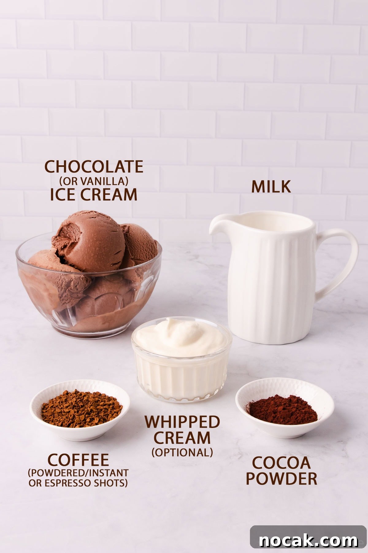 Creamy Mocha Milkshake Recipe: How to Make the Perfect Coffee Chocolate Shake 4 The mocha milkshake ingredients - chocolate ice cream in a glass bowl, milk in a white jug, coffee, cocoa powder and whipped cream in small bowls, on a pale grey marble background.