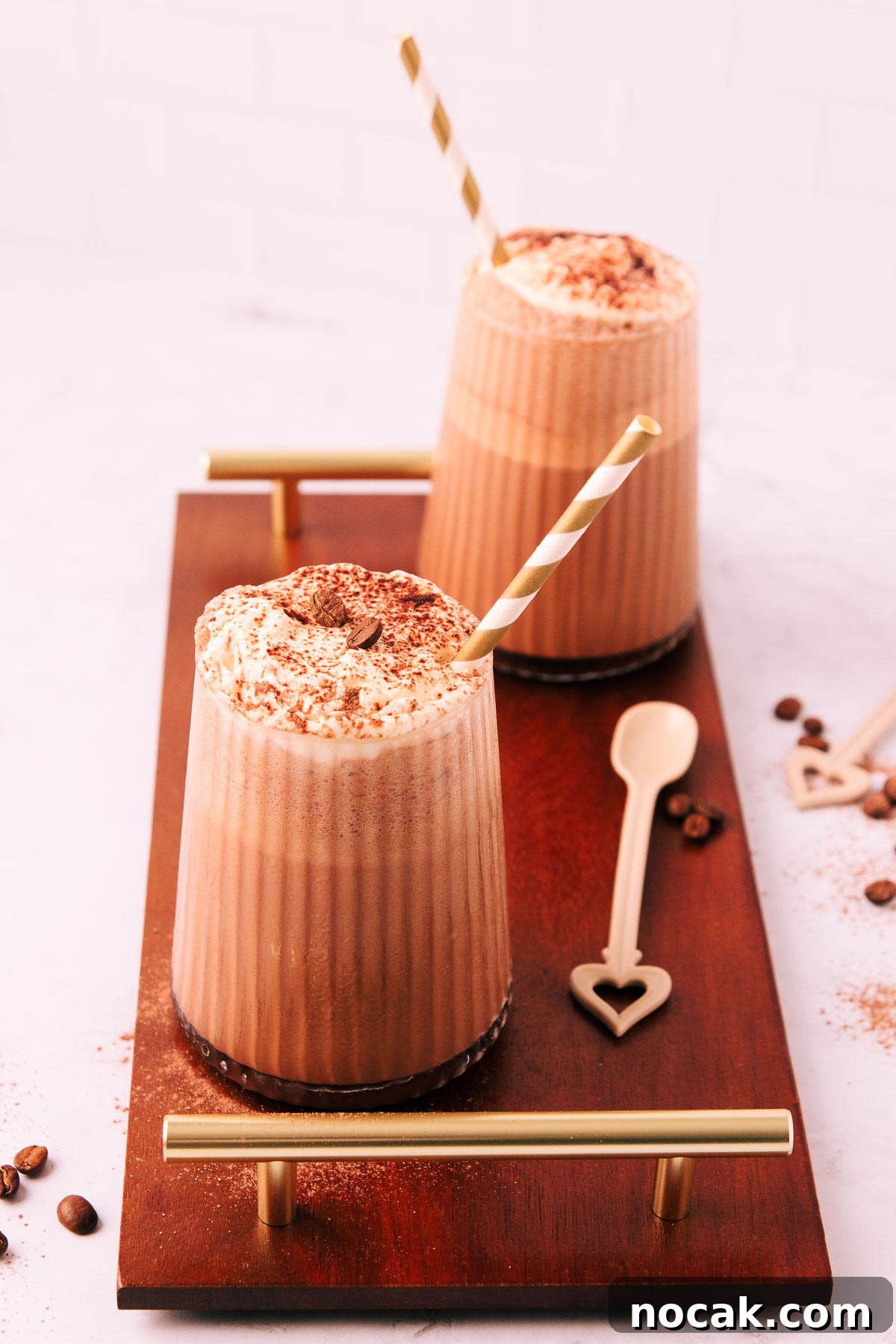 Creamy Mocha Milkshake Recipe: How to Make the Perfect Coffee Chocolate Shake 7 Two mocha milkshakes in glasses with a ribbed texture, topped with whipped cream and a sprinkle of cocoa, with gold and white striped paper straws on a dark wooden board with gold handles.