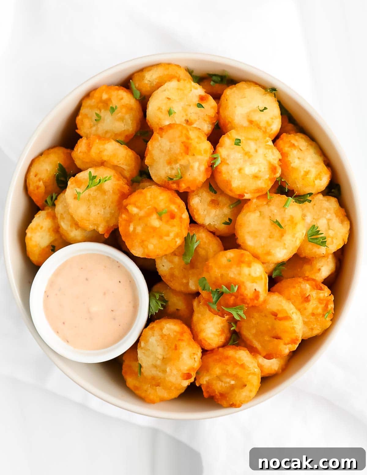 A white bowl filled with cooked tater tots and a small bowl of spicy ranch dressing. There is fresh parsley on top.