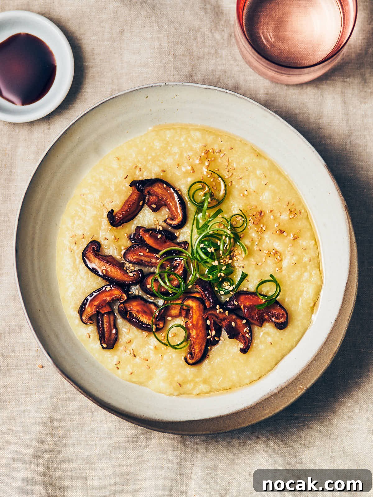 A bowl of congee with sauteed mushrooms, toasted sesame oil, sesame seeds, and scallions on top with a small dish of soy sauce on the side.