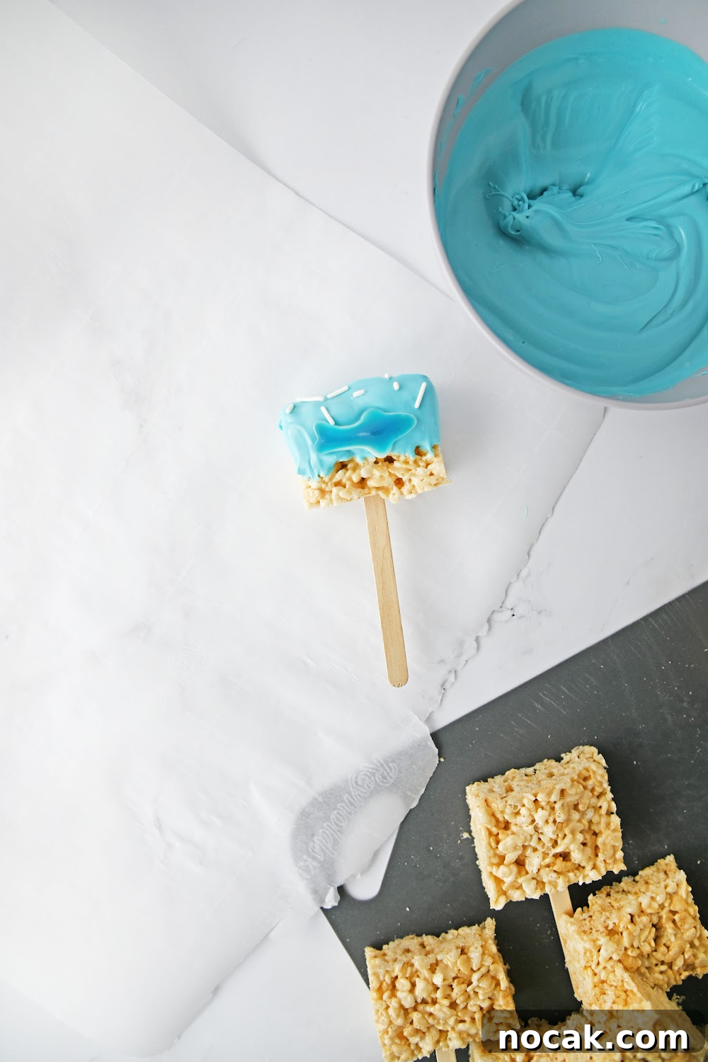 Shark-Themed Rice Krispie Treats: Fun Party Dessert Recipe 5 1 shark Rice Krispie treat pops made with a gummy shark laying on parchment paper with undecorated rice Krispie treats.