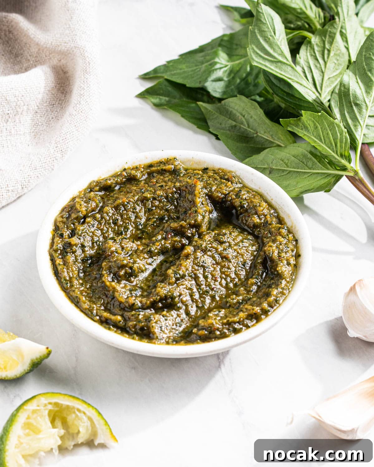 Side angle to show texture of bowl of Thai Basil Pesto with Thai basil, garlic cloves, and squeezed lime wedges surrounding.
