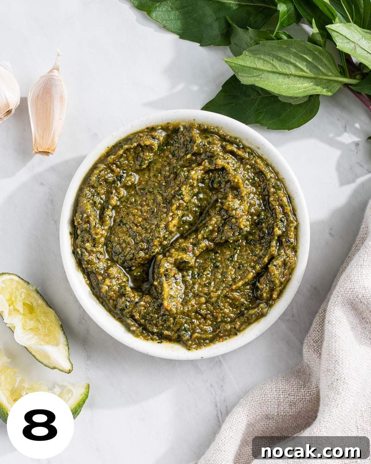 Step 8: Bowl of Thai Basil Pesto with Thai basil, garlic cloves, and squeezed lime wedges surrounding.
