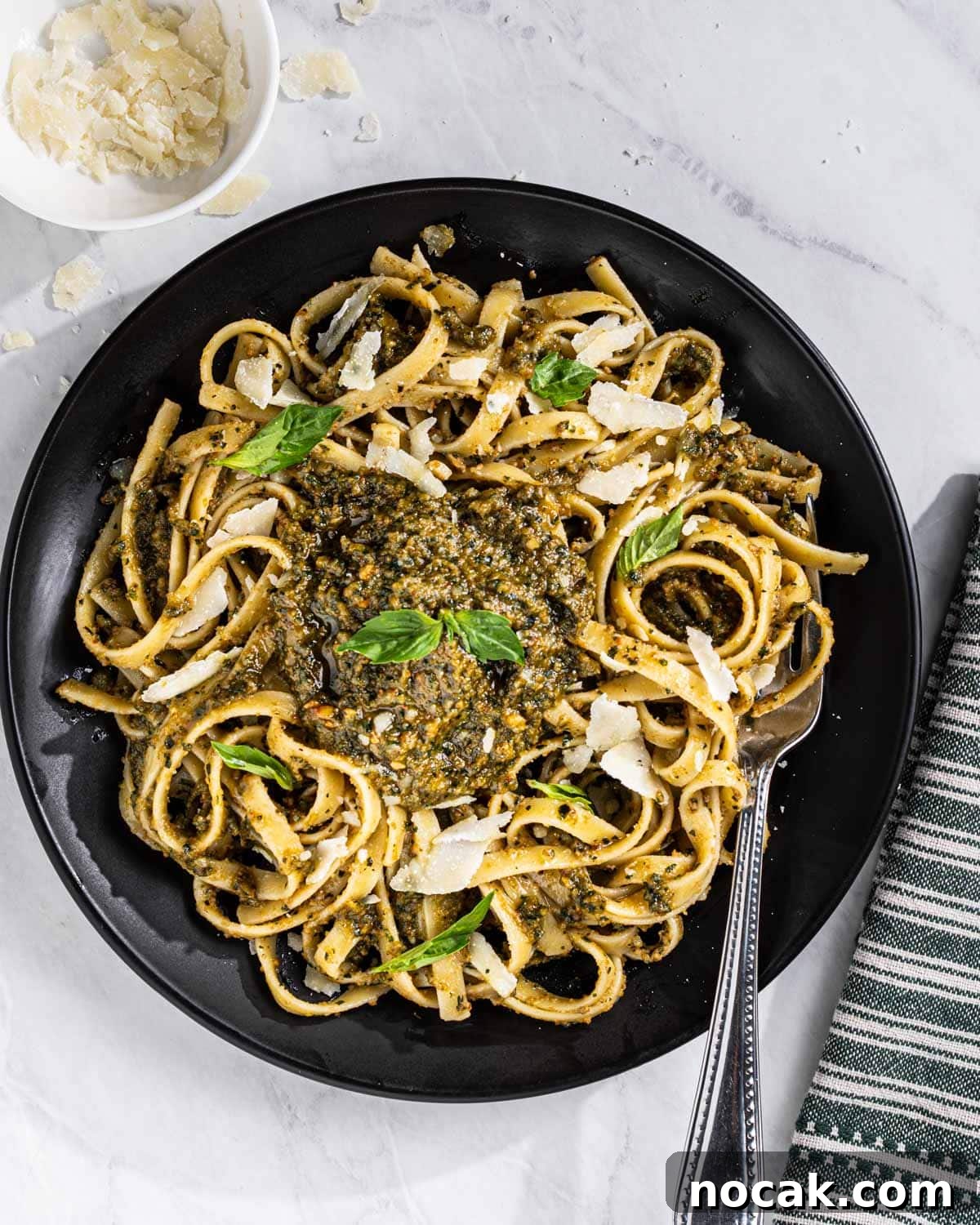 Black Plate with Thai Basil Pesto Pasta topped with a dollop of Thai Basil Pesto, shaved Parmesan cheese, and Thai Basil leaves. Fork and tea towel on the side.