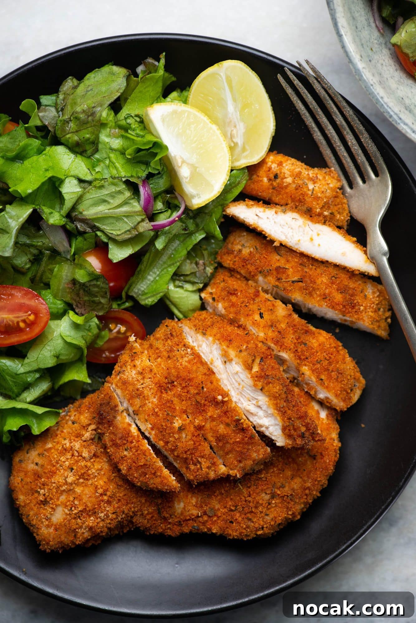 Crispy Air Fryer Chicken Breast 7 Sliced crispy crusty chicken breasts served with chopped lettuce, cherry tomatoes and lemon on a black plate.