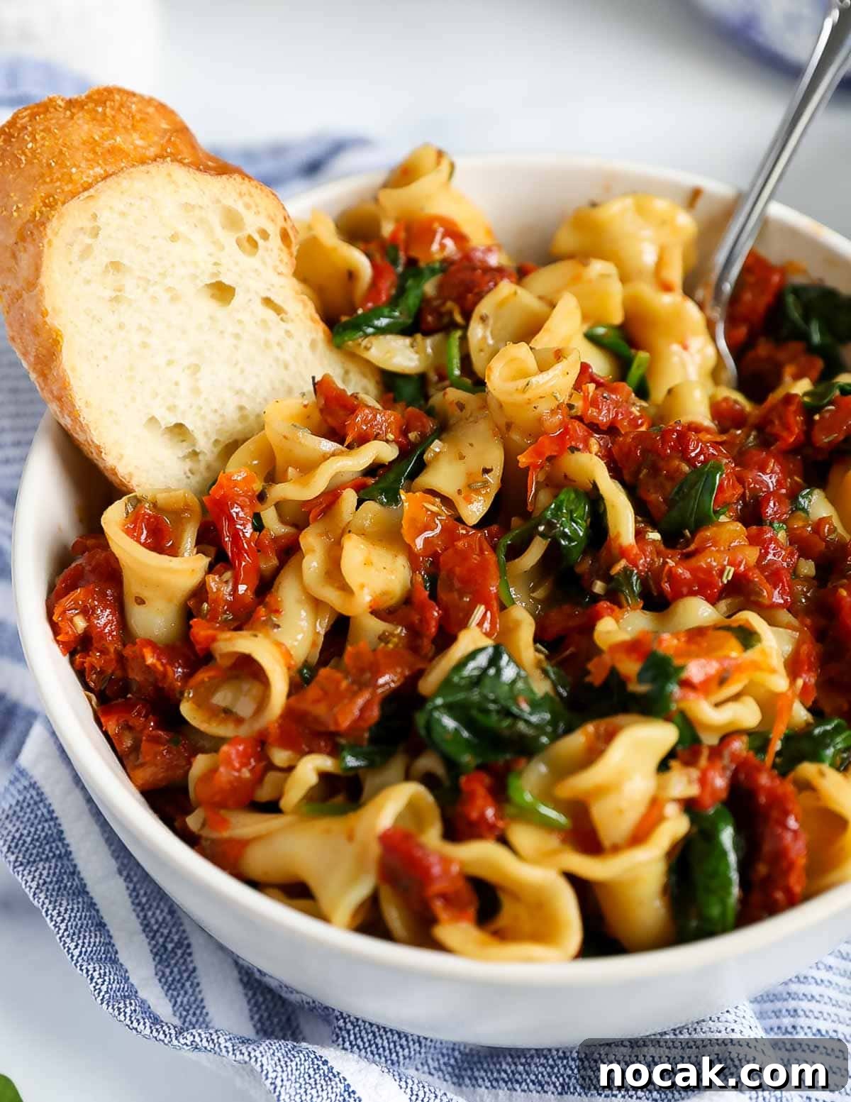Creamy Tuscan Garlic Pasta with Sun Dried Tomatoes 2 A fork placed inside a white bowl that is filled with pasta, tomatoes, and spinach. There is also a slice of bread inside the bowl.